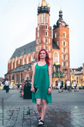 girl with red hair alone in a green dress holds a smartphone in her hands. Reflection in a puddle. Travel and tourism concept, vertical photo. central square of Krakow, Poland