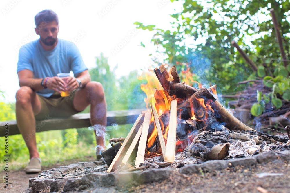 Man drinking beer while sitting on bench at park with bonfire Man drinking beer while sitting on bench at park with bonfire
