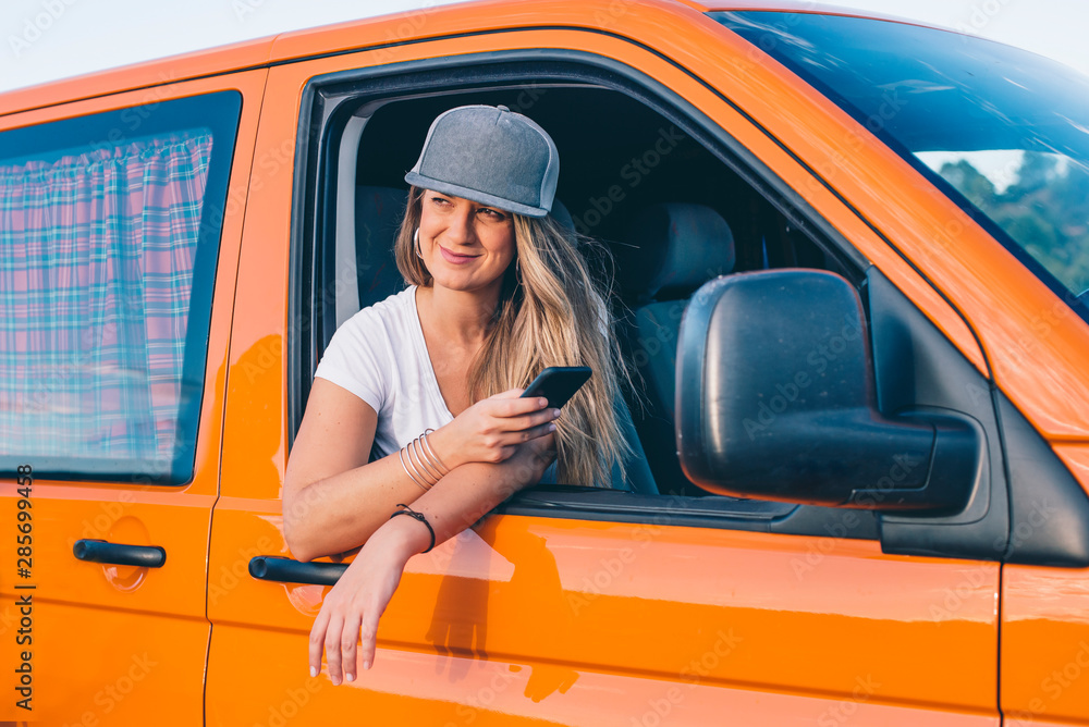 Naklejka premium pretty and attractive blonde girl with cap wearing phone in orange van with mountains in the background and gray sky