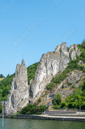 Dinant, Belgium - June 26, 2019: South side of Le rocher Bayard along La Meuse River surrounded by green foliage under blue sky.