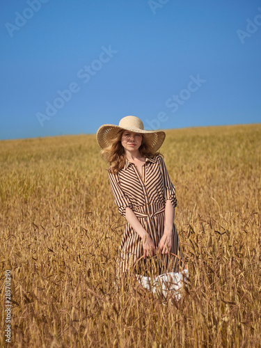 A blonde in a wheat field walks with a wicker basket of apples in a lime hat and a vertical striped dress.