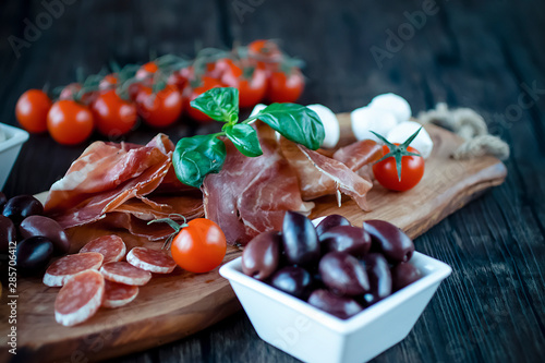 Prosciutto, bread, olives, walnut, mozzarella, salami, basil and cherry tomatoes on brown wooden board. Mediterranean kitchen. Top view.