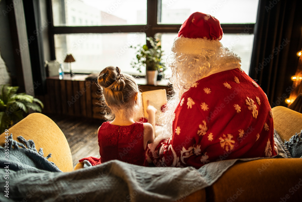 Obraz premium Santa Claus with a child reads a fairy tale against the window where the Christmas tale begins