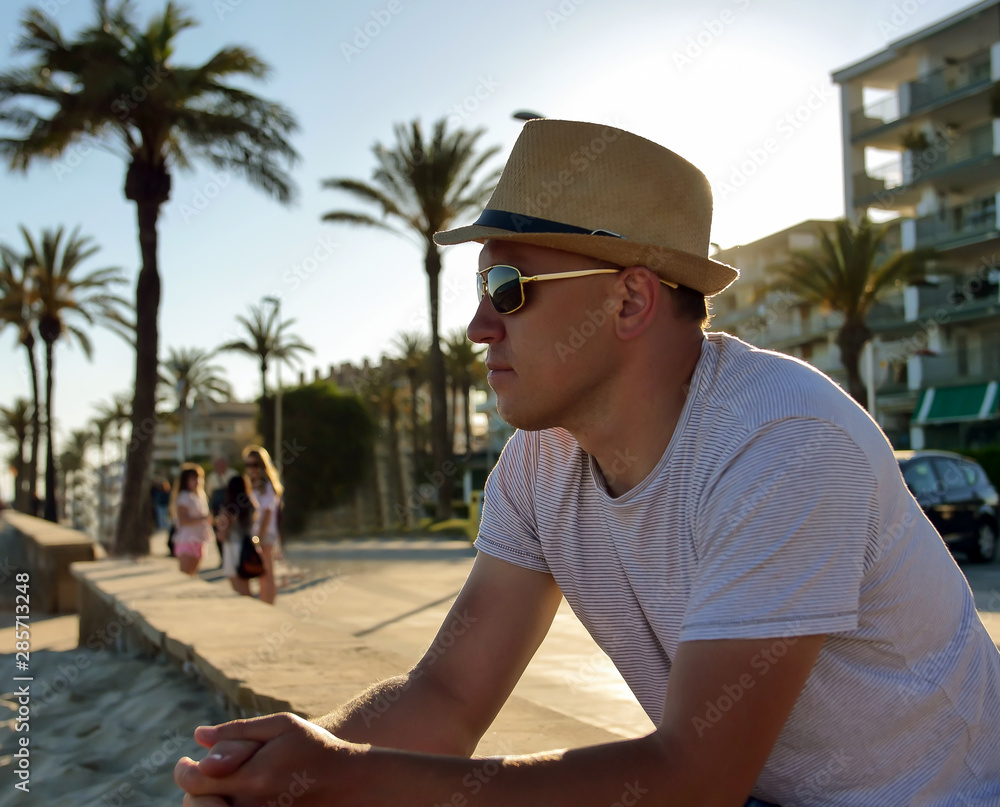 Young attractive man in a hat and sunglasses sits on the waterfront by the sea