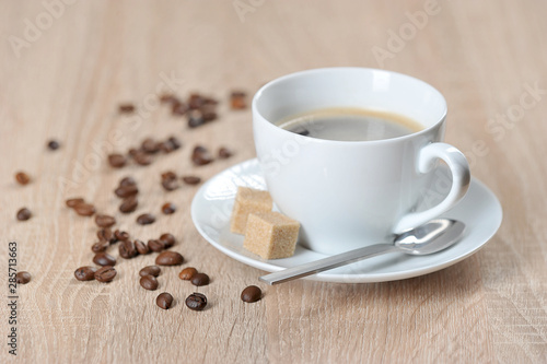 Coffee in a white cup and saucer. On a saucer are two pieces of cane sugar and a teaspoon. Coffee beans on the surface of the table. Light background. Free space for text.