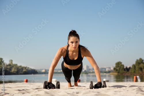 Young healthy woman training upper body with weights at the beach. Single caucasian female model practicing at the river side in sunny day. Concept of healthy lifestyle, sport, fitness, bodybuilding.