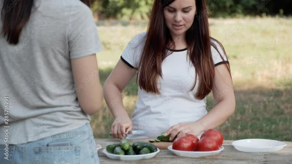 Two young women at a wooden table in the middle of nature, a pine forest, chatting among themselves preparing a salad of fresh tomatoes and cucumbers. Two young women at a wooden table in the middle of nature, a pine forest, chatting among themselves preparing a salad of fresh tomatoes and cucumbers.