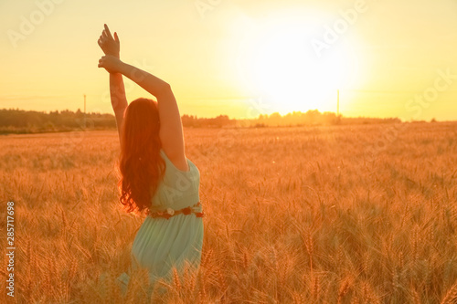 girl in dress walking in golden ripe wheat field at sunset with hands upwith  photo