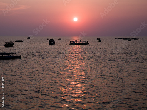 Sunset over Lake Tonle Sap. Several boats spread over the water close to horizon line. Sun reflecting on the water surface. Golden hour picture - various tones of purple colour. 