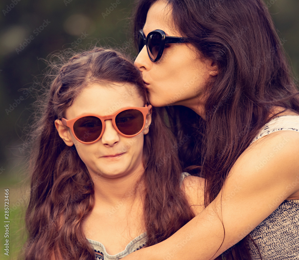 Happy beautiful woman kissing and embracing her fun grimacing daughter in trendy sunglasses on nature summer background. Closeup