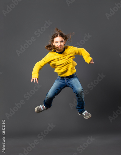 Funny happy child girl jumping in casual blue jeans and yellow pullover on grey studio background