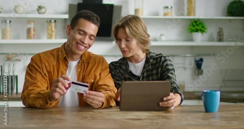 Happy young mixed race couple is doing online shopping, using credit card and tablet computer, enjoying shopping time, sitting at table in cozy kitchen, Hispanic, Caucasian. 4K, shot on RED camera.