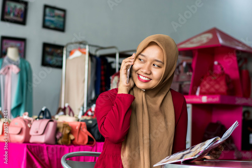 portrait of a young muslim entrepeneur sitting and smile talking in her smartphone in front of fashion boutique as background -image
