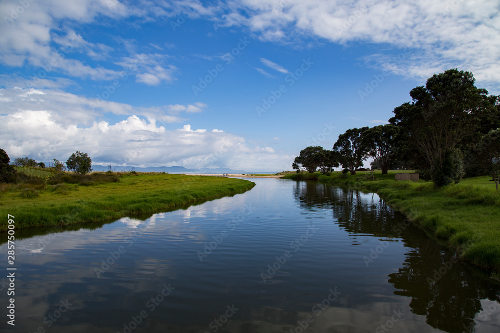 Stunning Kuaotunu estuary in Coromandel on picture perfect summers day. Blue sky, clouds, reflections on water. Native trees pohutukawa and flax. NZ golden sand, blue sea. One of best places on earth.