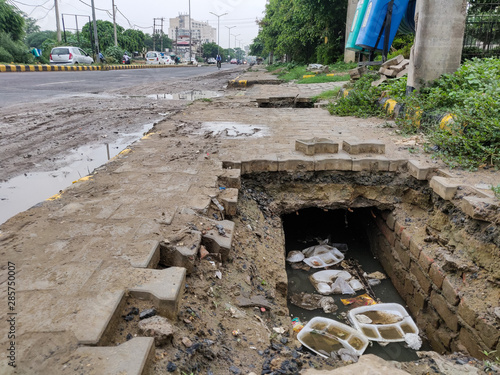 Billede på lærred Broken footpath exposes underground sewage gutter filled with rain water, garbage, plastic waste, food trays