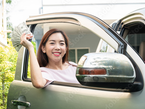Asian woman holding money while standing beside a car, insurance, loan and finance concept