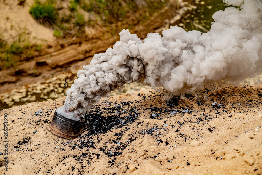 Fototapeta premium Grey smoke comes out from smoke bomb. The bomb on sand near a forest. Military training. Air pollution