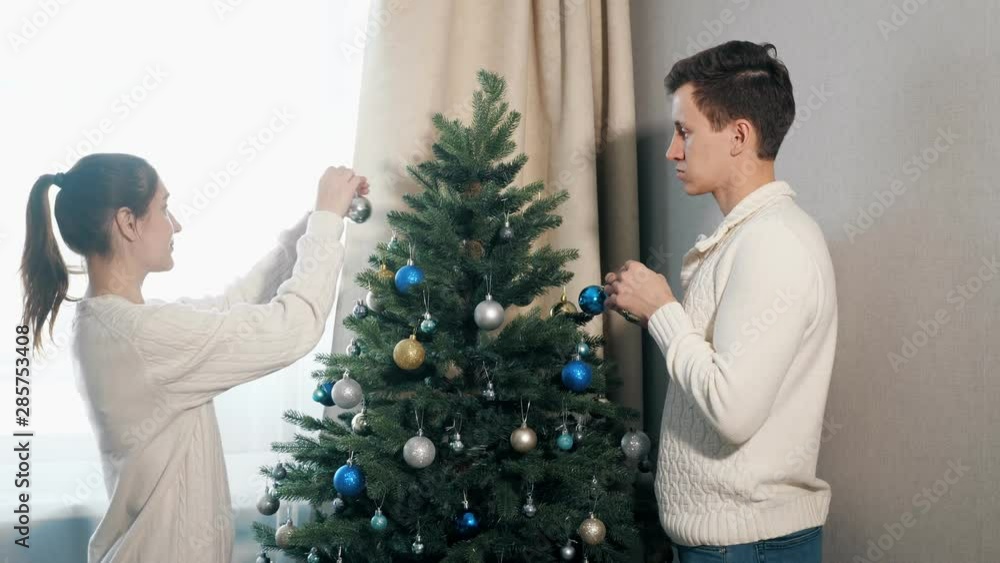 handsome young man and pretty lady in white sweaters hold colorful toys decorating artificial christmas tree close view