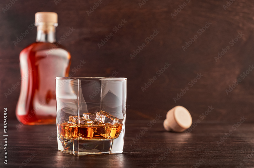 A glass of whisky with ice on a wooden table, a full bottle of whisky in the background A glass of whisky with ice on a wooden table, a full bottle of whisky in the background