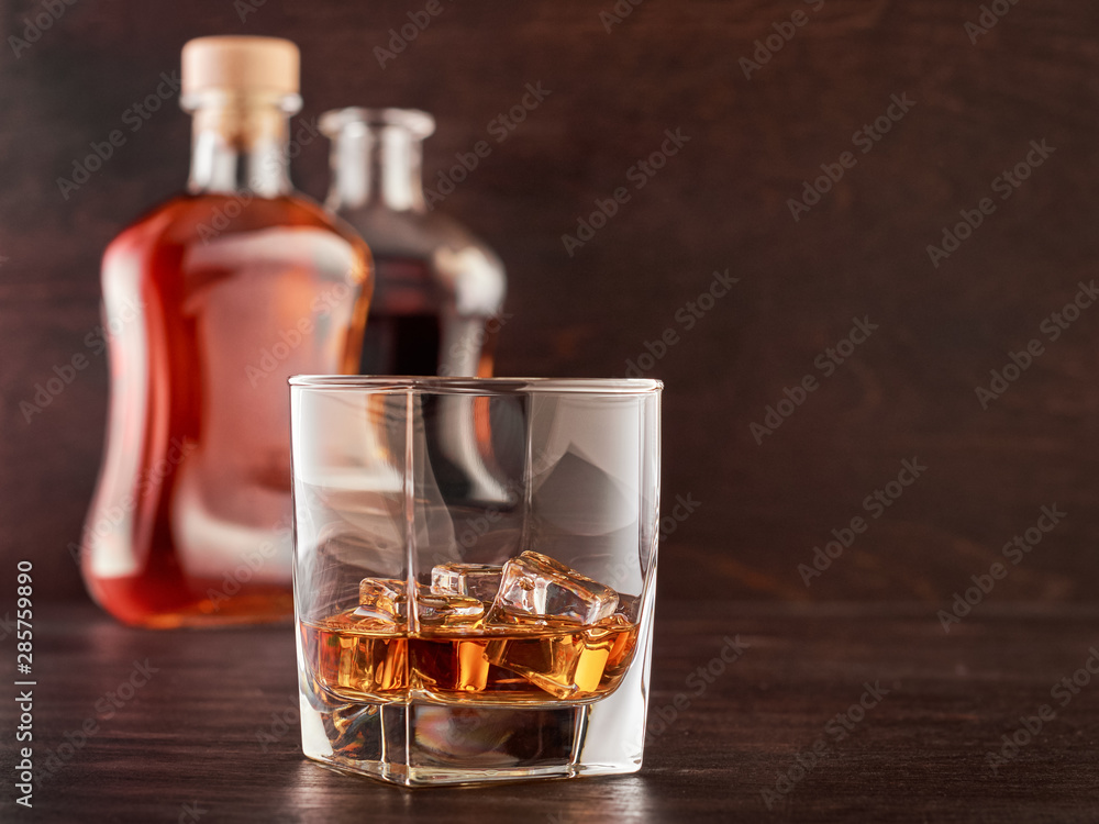 A glass of whisky with ice on a wooden table, two bottles of whisky in the background, full and open A glass of whisky with ice on a wooden table, two bottles of whisky in the background, full and open