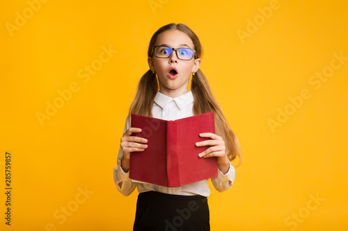 Funny Shocked Schoolgirl Posing With Book On Yellow Background