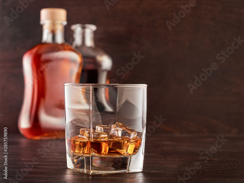 A glass of whisky with ice on a wooden table, two bottles of whisky in the background, full and open