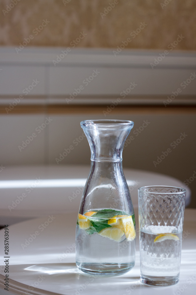 Close-up of a bottle with a cap and a glass of water with mint and lemonon a white table and sun glare, selective focus