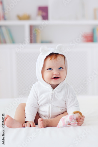 Murais de parede happy infant baby girl in funny ear hood is sitting on the bed at home
