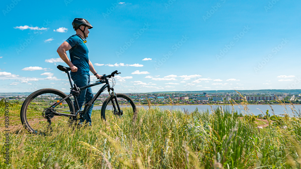 Obraz premium A male cyclist stands on a hill and looks at the city. A man in a helmet and with a bicycle on nature with his back to the camera.