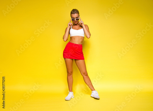 young sports happy sexy blonde girl in red skirt and white top is posing like model and looking down with sunglasses in her hands on the yellow wall background, sport lifestyle concept, free space
