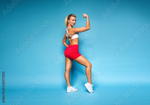 young sports happy sexy blonde girl in red skirt and white top is standing like athlete with one strong hand, looking smily and flirting on blue wall background, sport lifestyle concept, free space