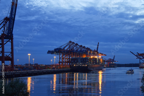 Illuminated Container Ship / Freighter at Dusk in the port of Hamburg with big Gantry Cranes (Container Cranes). Harbor Tour Boat next to it