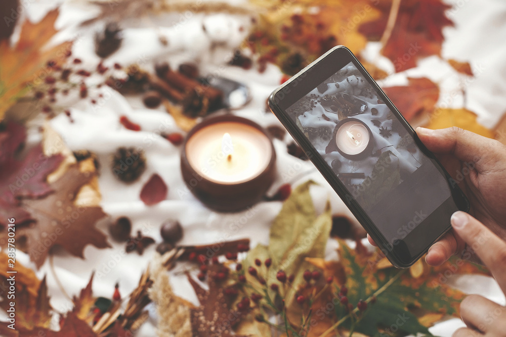 Autumn hygge flat lay. Hand holding phone and taking photo of candle, berries, fall leaves, anise,herbs, acorns, nuts, cinnamon, cotton on white textile. Hygge lifestyle, cozy autumn mood Autumn hygge flat lay. Hand holding phone and taking photo of candle, berries, fall leaves, anise,herbs, acorns, nuts, cinnamon, cotton on white textile. Hygge lifestyle, cozy autumn mood