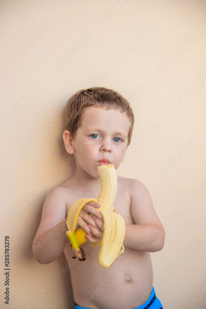 The boy eats a banana. The boy on the background of a plain wall. Fruits . The boy eats a banana. The boy on the background of a plain wall. Fruits .