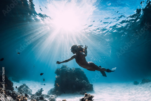 Woman freediver with fins swim over sandy sea with fish and sun rays underwater