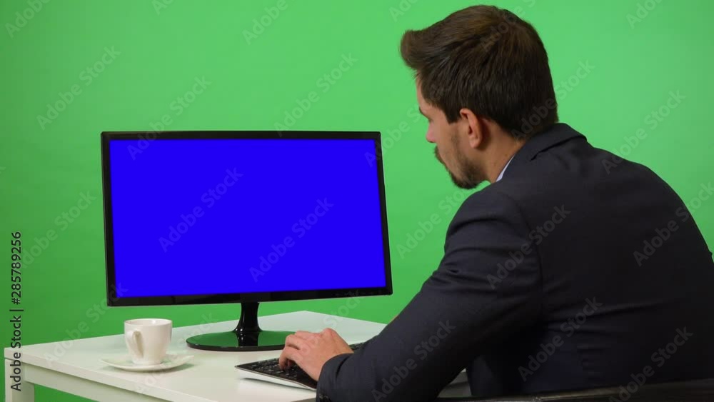 A young businessman sits at a desk and works on a desktop computer with blue screen - green screen studio - he types on keyboard A young businessman sits at a desk and works on a desktop computer with blue screen - green screen studio - he types on keyboard