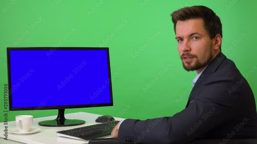 A young businessman sits in front of a computer with blue screen and talks seriously to the camera - green screen studio A young businessman sits in front of a computer with blue screen and talks seriously to the camera - green screen studio