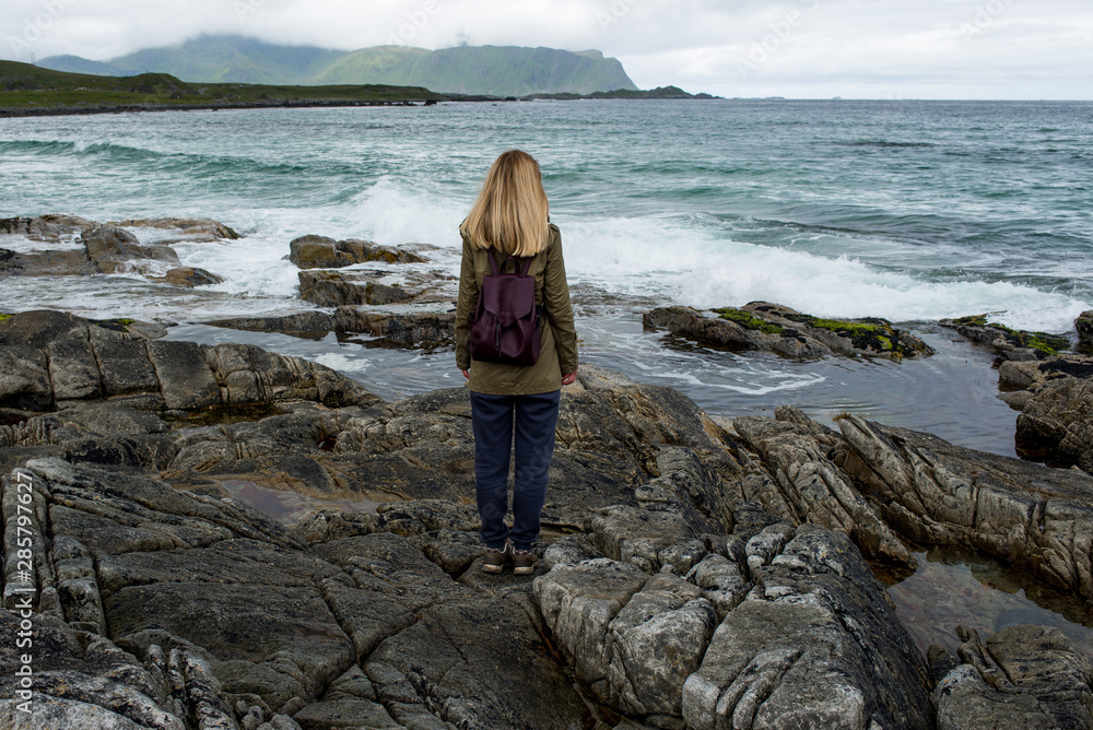 Blond hair girl with a backpack stands on big stones near the water and looks at the ocean. Waves, splashing. Enjoy the moment, relaxation. Wanderlust. Travel, adventure, lifestyle. Explore Norway