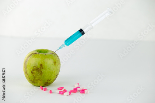 Concept: human GMO manipulation of nature and relative poisoned fruits. Close-up of an apple contaminated with a syringe threaded in and medicines on a white background