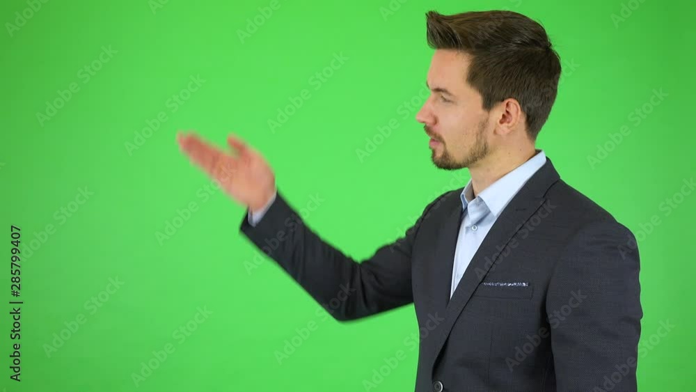 A young handsome businessman talks to the camera and gestures toward a list next to himself, runs his hand from top to bottom - green screen studio A young handsome businessman talks to the camera and gestures toward a list next to himself, runs his hand from top to bottom - green screen studio