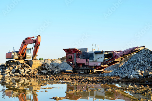 Heavy excavator working in quarry on a background of sunset and blue sky. Mobile jaw stone crusher by the construction site. Crushing old concrete wastes and subsequent cement production