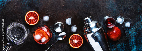 Negroni alcoholic cocktail beverage with dry gin, red vermouth and red bitter, bloody orange slice and ice cubes. Brown bar counter background, steel bar tools, top view, with copy space