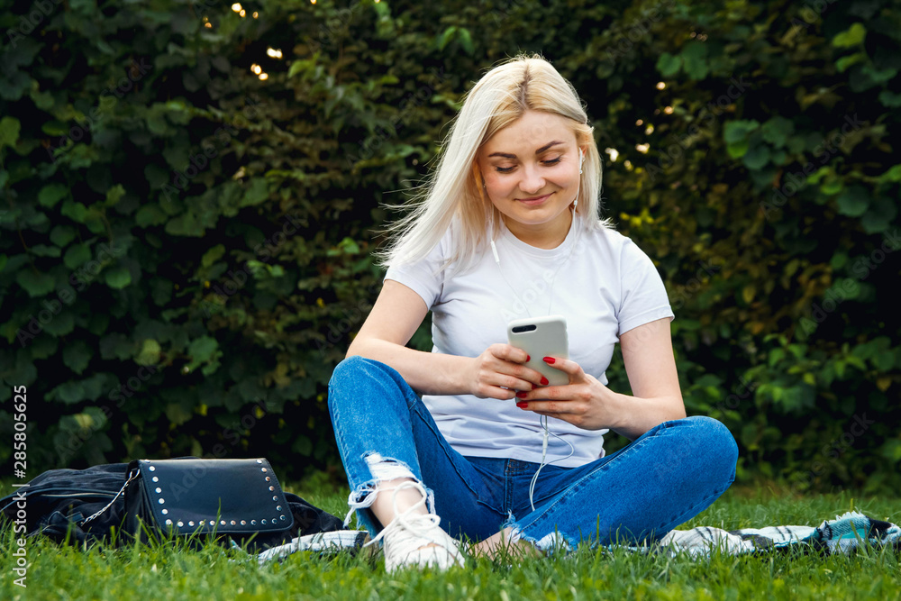 Beautiful blonde girl in headphones and smartphone in hands sits on green grass. She is talking on the phone through headphones. Lifestyle concept. Copy, empty space for text Beautiful blonde girl in headphones and smartphone in hands sits on green grass. She is talking on the phone through headphones. Lifestyle concept. Copy, empty space for text