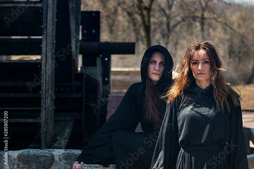 Young Women with dark long hair in black robes in front of an Old Wooden Water Mill. Witches. Halloween concept. Witchcraft and Magic. 