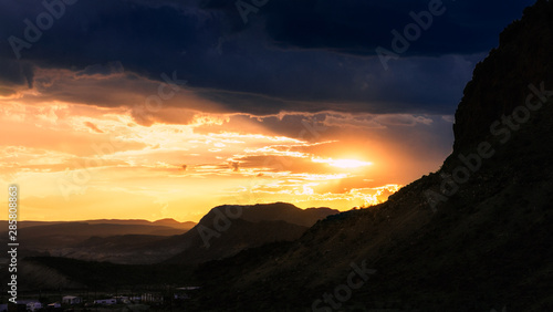 The setting sun casts an orange glow over the mountain landscape as dark storm clouds fill the sky