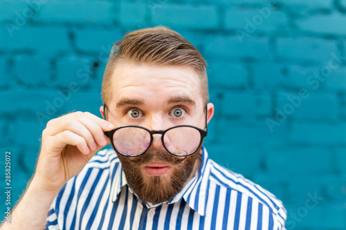 Close-up portrait of a curious surprised young man in glasses with a mustache and beard posing on a background of blue blurred brick wall. Concept of surprise and shocking information.