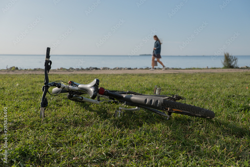 Report from the seashore. The bicycle lies on a green grass. People walk along the water. Summer. Sunny day.