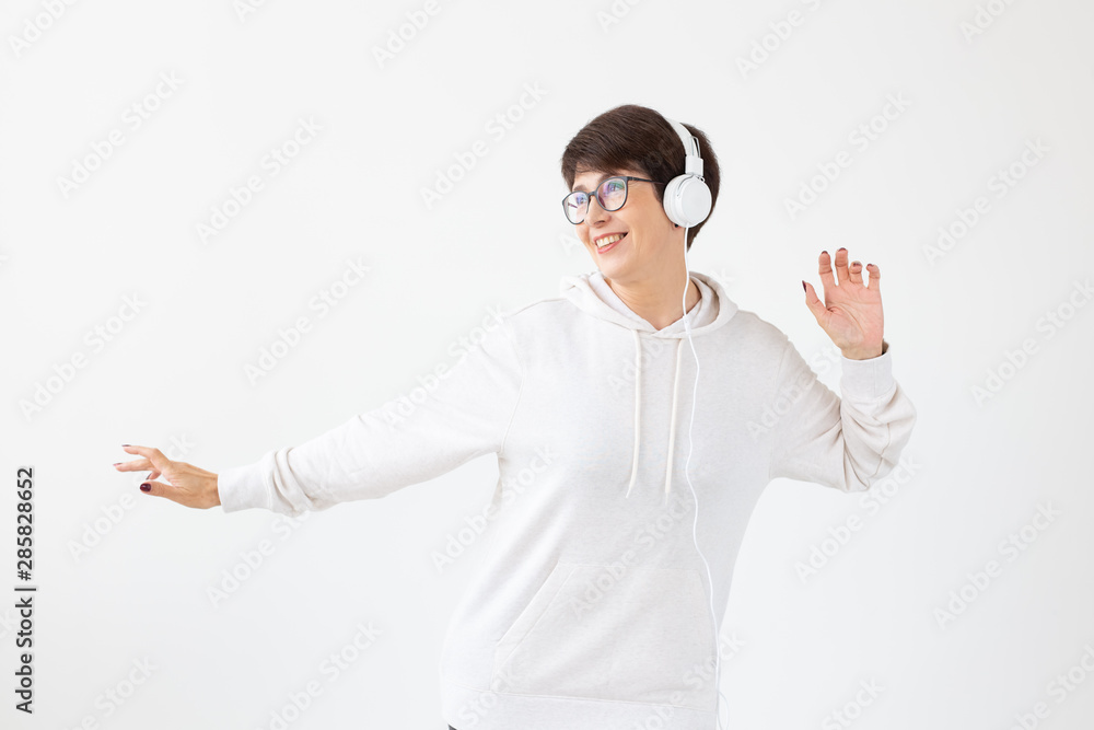 Cute positive middle-aged woman in sweater and glasses is listening to music with wire headphones standing on a white background. Concept of hobbies and subscriptions to favorite radio station. Cute positive middle-aged woman in sweater and glasses is listening to music with wire headphones standing on a white background. Concept of hobbies and subscriptions to favorite radio station.
