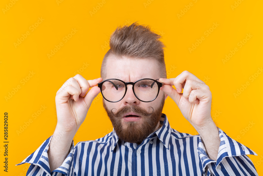 Young handsome guy is looking at his glasses while standing against a yellow background. The concept of poor vision and eye treatment.