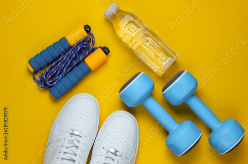 Flat lay style sport concept. Dumbbells, sneakers, jump rope, bottle of water. Sports equipment on yellow background. Top view